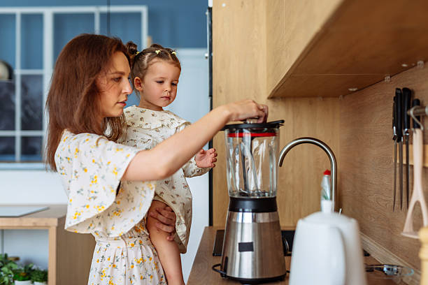 Mother use blender to prepare food with small child in her arms